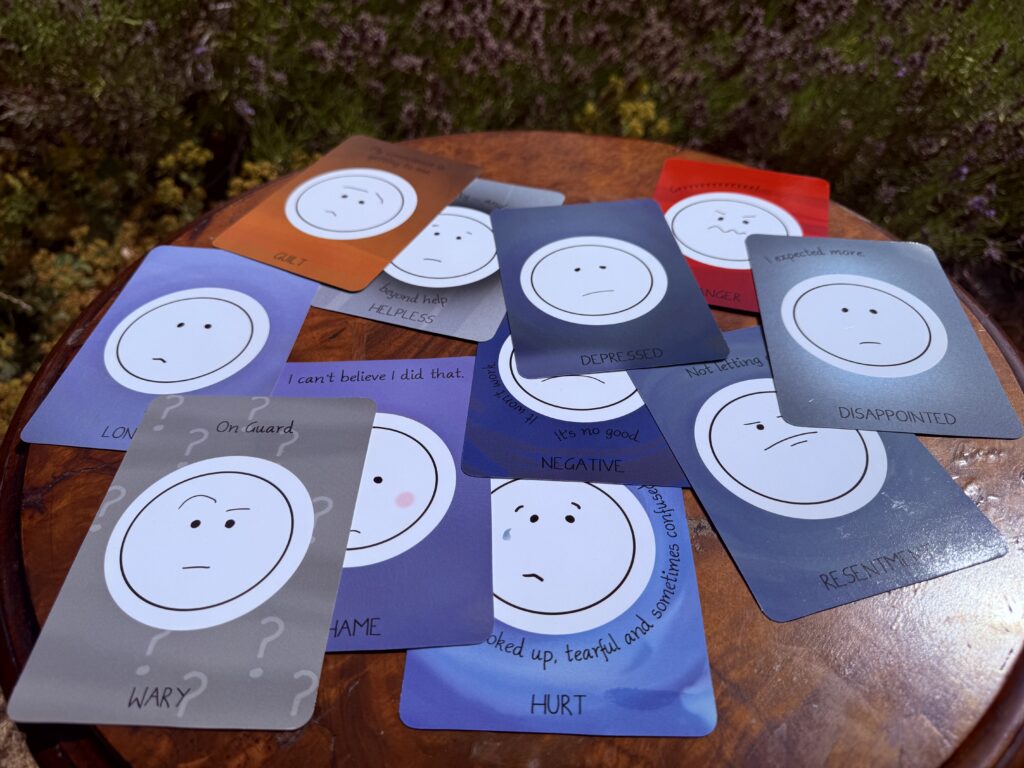 A selection of cards all showing unhappy or angry faces representing different emotions spread across a round wooden table in front of green plants.