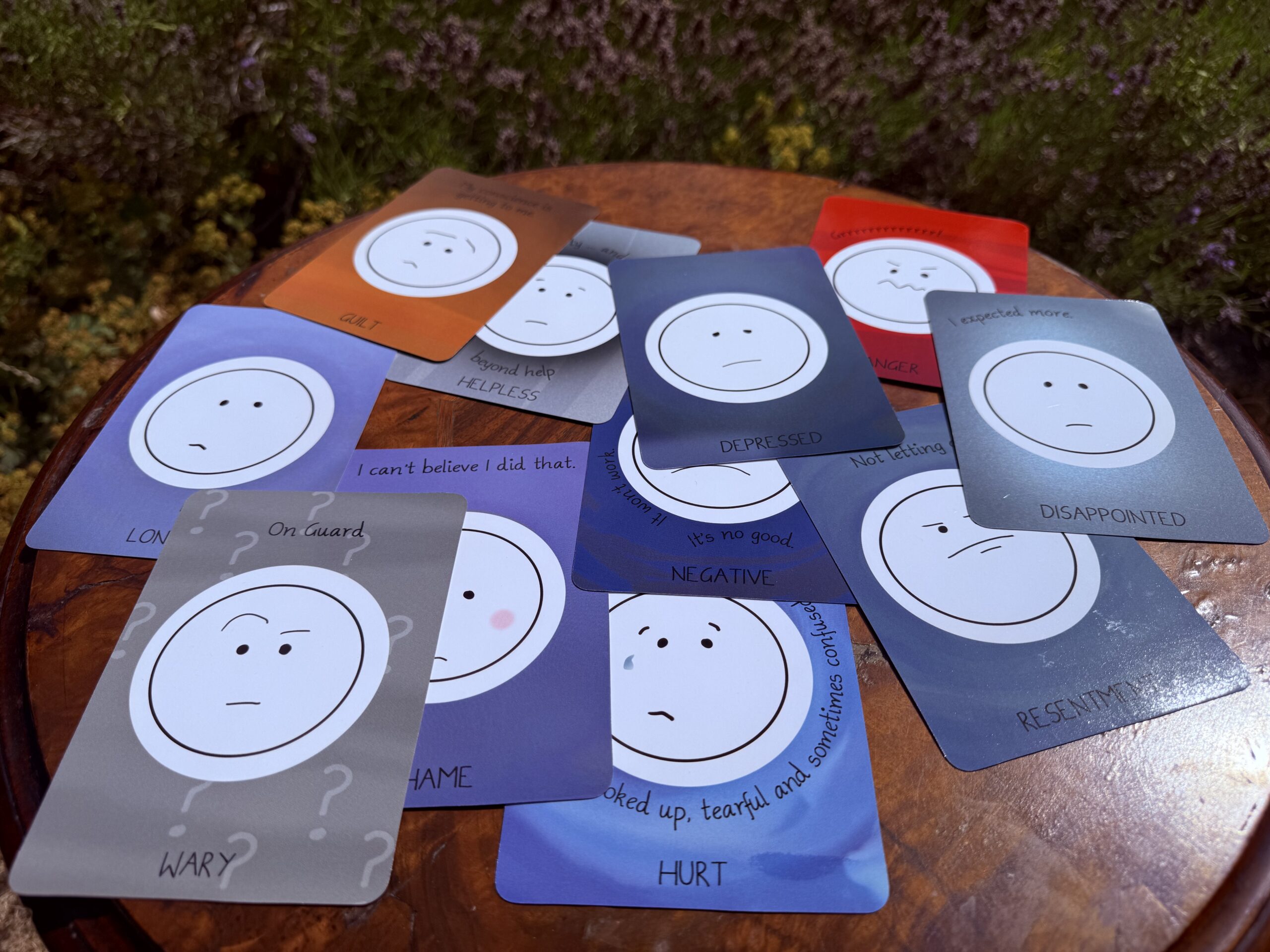 A selection of cards all showing unhappy or angry faces representing different emotions spread across a round wooden table in front of green plants.