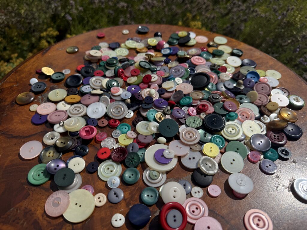 A large selection of different colourful buttons spread across a round wooden table in front of green plants.